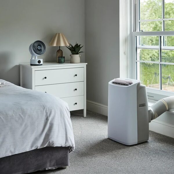 A white portable air conditioner with an exhaust hose vented out of a sash window in a grey bedroom