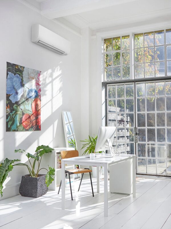 A white wall-mounted air conditioning unit installed high on the wall of a bright, airy home office with a desk, plants and large windows