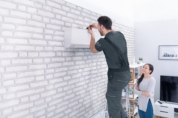 A technician on a ladder installing a wall-mounted air conditioning unit in a living room while a woman watches