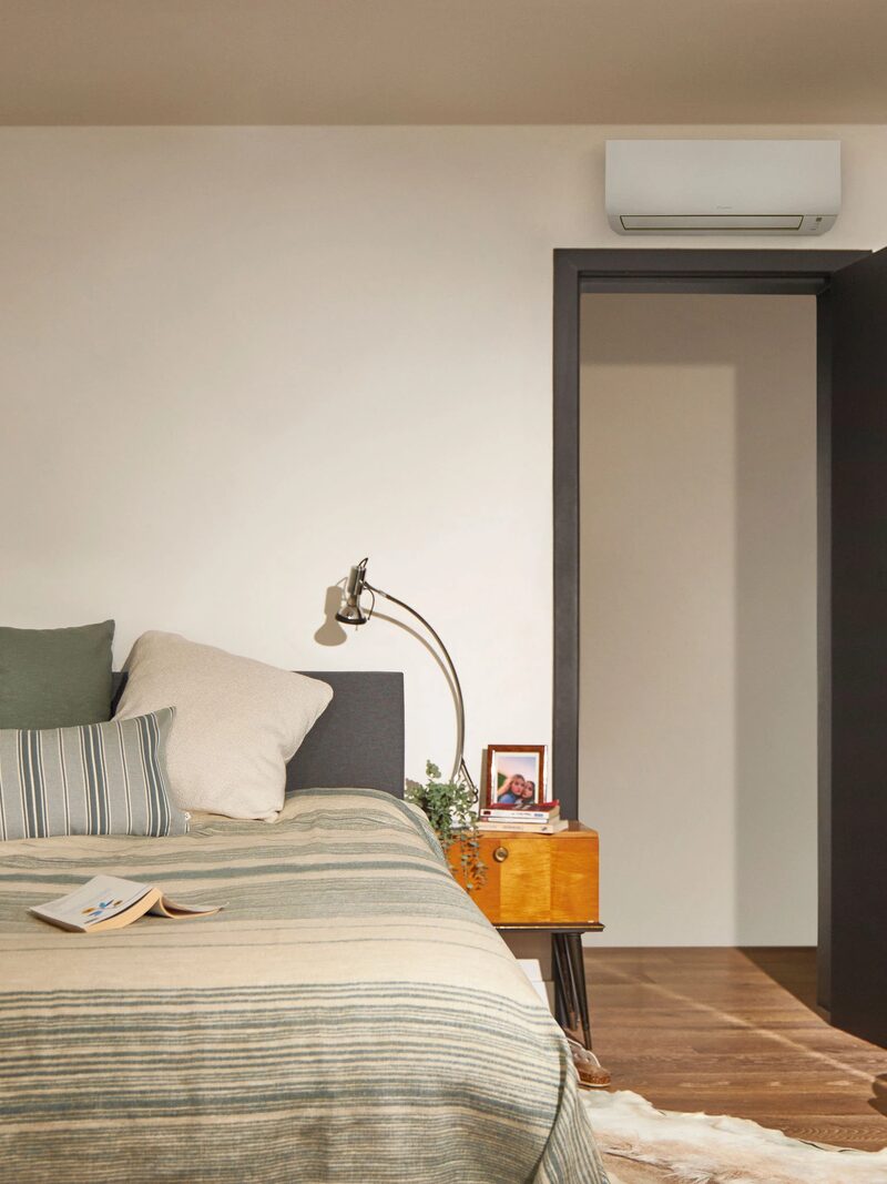 A wall-mounted air-source heat pump indoor unit above a doorway in a cosy bedroom with a made bed