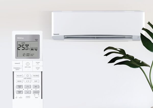 A white Panasonic wall-mounted air conditioning unit with its remote control in front of a pale wall beside a houseplant