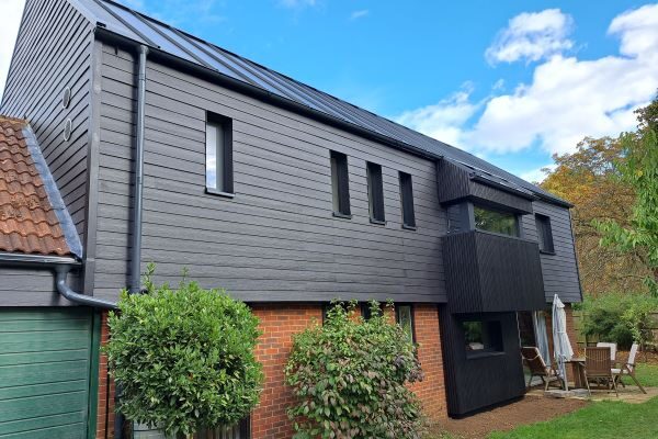 A dark timber-clad Passivhaus retrofit house with a metal roof and solar panels, seen from the garden