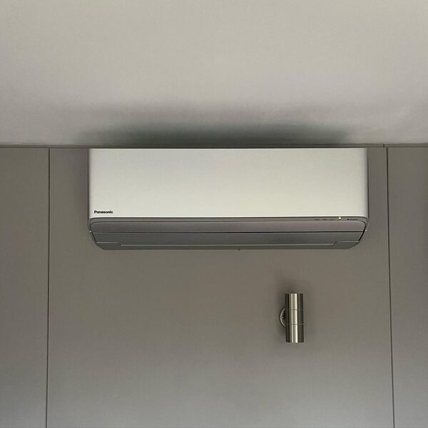 A white Panasonic wall-mounted air conditioning unit installed high on a grey interior wall next to a small wall light