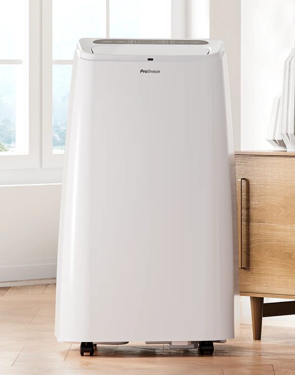 A white Pro Breeze portable air conditioner standing on castors in a bright room beside a wooden sideboard