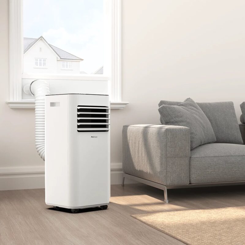 A white Pro Breeze portable air conditioner with an exhaust hose vented out of a window in a bright living room beside a grey sofa