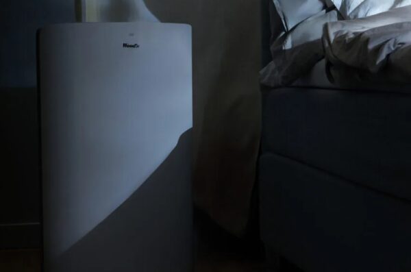 A white Woods portable air conditioner standing in a dim bedroom next to a bed