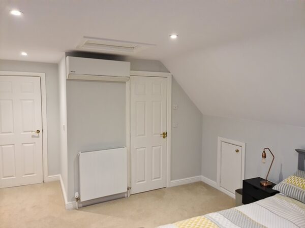 A wall-mounted Panasonic air conditioning unit installed high on the wall of a loft-conversion bedroom, with a radiator below