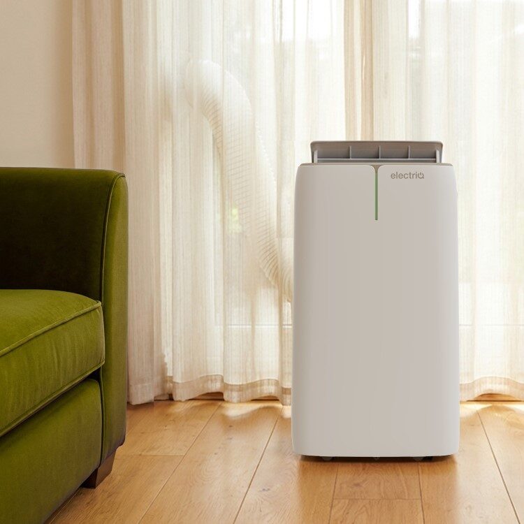 A white electriQ portable air conditioner with exhaust hose standing on a wooden floor next to a green sofa and sheer curtains