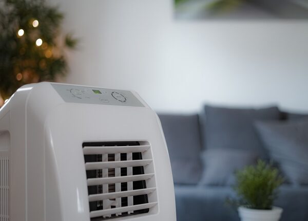 A white portable air conditioning unit standing in a living room with a grey sofa in the background