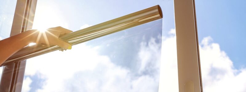 A hand applying reflective solar-control film to a window pane with blue sky and sun behind