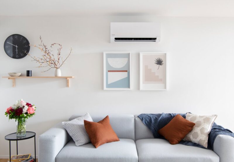 A white wall-mounted split air conditioning unit installed above a grey sofa in a bright living room