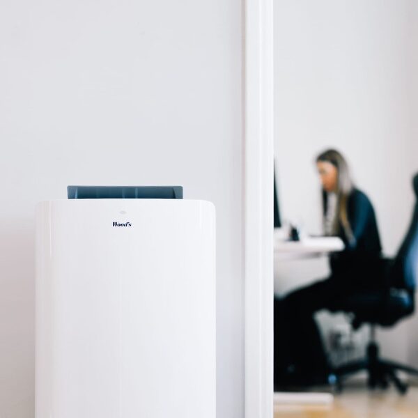 A white Wood's portable air conditioner standing in a room with a woman working at a desk in the background