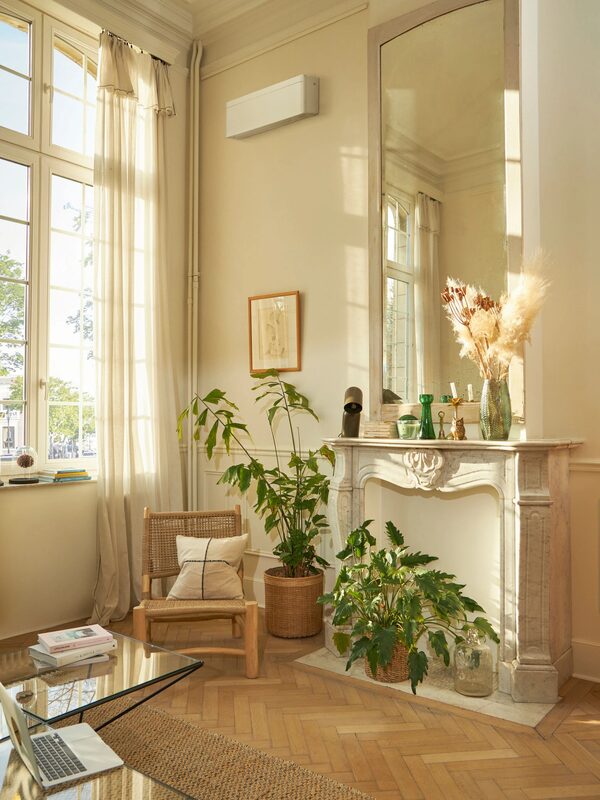 A white wall-mounted air conditioning/heat pump indoor unit installed high on the wall of an elegant cream-coloured living room with a marble fireplace, mirror and plants