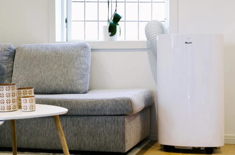 A white Woods portable air conditioner standing in a living room next to a grey sofa, with its exhaust hose running to the window
