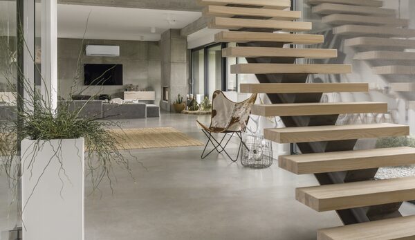 A wall-mounted air conditioning unit high on the wall of a modern open-plan living room with a floating wooden staircase