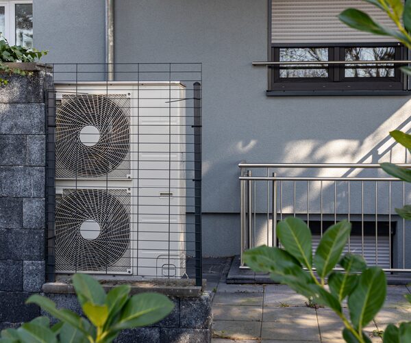 An air source heat pump with two fans installed outside a grey house behind a wire guard