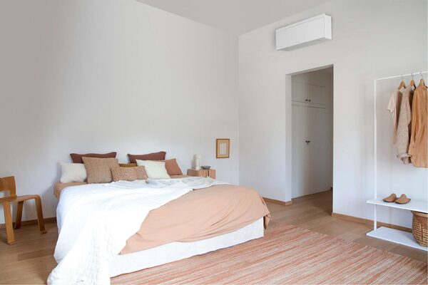 A white wall-mounted air conditioning unit installed high on the wall of a bright minimalist bedroom