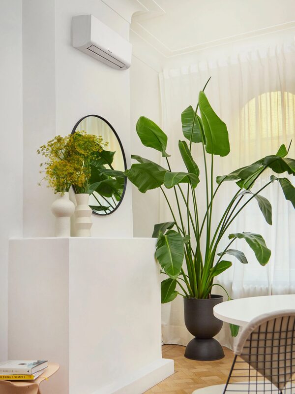 A white wall-mounted air conditioning unit installed high on a wall in a bright living room with plants and a mirror