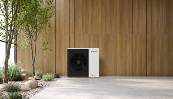 A Hitachi air source heat pump unit installed outdoors against a wooden panel wall beside a paved area with plants and rocks
