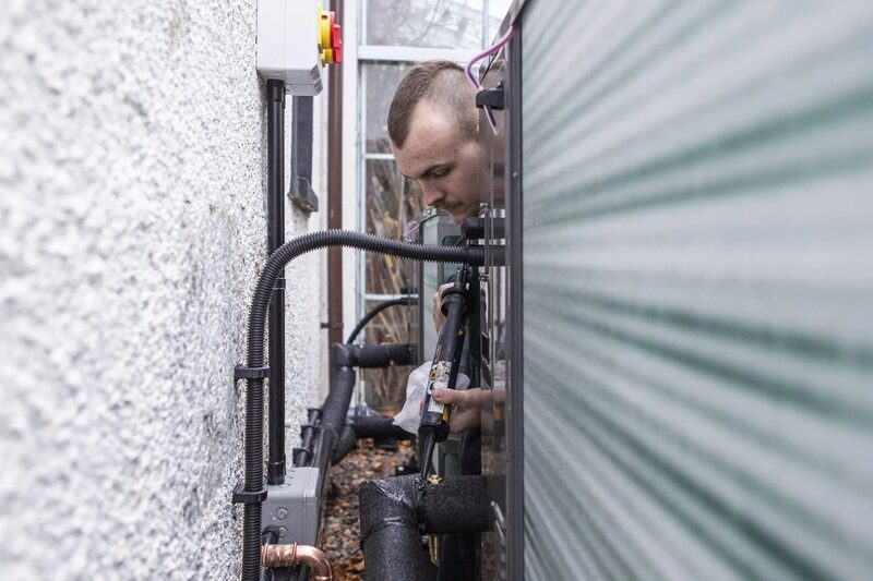 An installer working on insulated pipework behind an outdoor air source heat pump unit on the exterior wall of a house