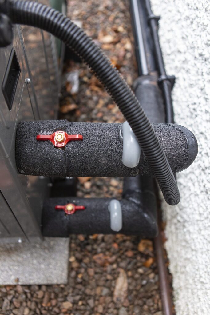 Insulated pipework with red isolating valves and black flexible conduit at the back of an outdoor heat pump unit