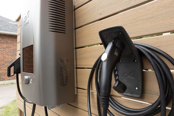 A dcbel Ara bidirectional EV charger unit mounted on a wooden slat wall outdoors with its charging cable and holster