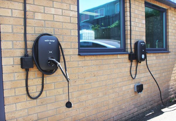 Two black waEV-charge EV chargers mounted on a brick wall outside a building with plugged-in cables