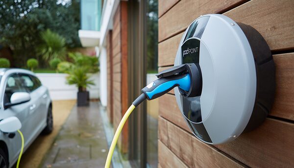 A Pod Point wall-mounted EV charger with a blue tethered cable plugged in, on a wooden-clad house wall beside a parked electric car