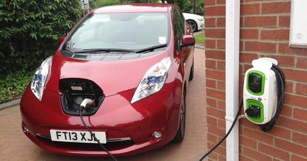 A red Nissan Leaf electric car charging from a wall-mounted home charge point on a brick house
