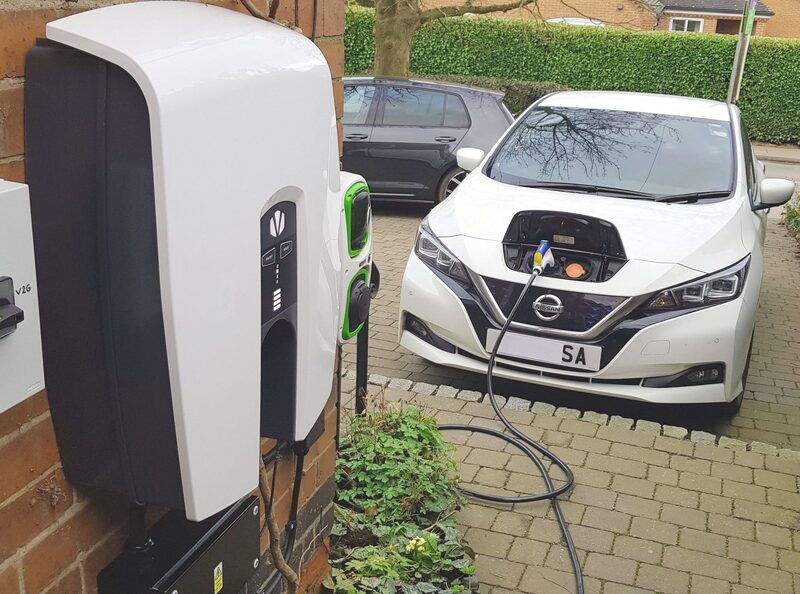 A white Nissan Leaf electric car charging from a wall-mounted EV charge point on a brick house driveway