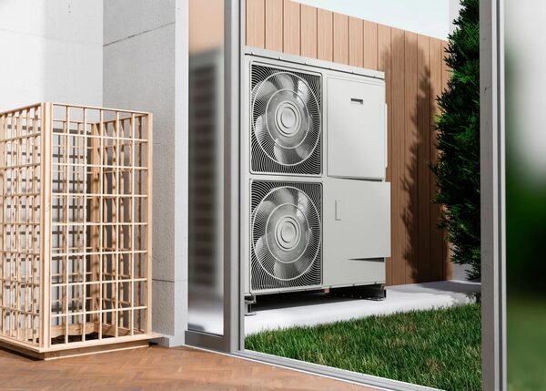 A large grey air source heat pump outdoor unit with two fans installed against a timber-clad wall beside a garden