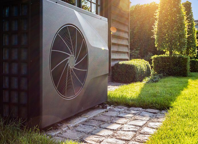 An air source heat pump unit installed outside a house on a cobbled path beside a sunny lawn