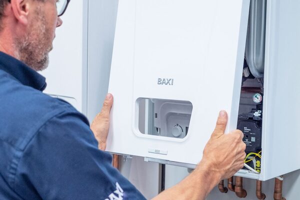A technician fitting the front casing onto a white Baxi wall-mounted combi boiler with copper pipework below