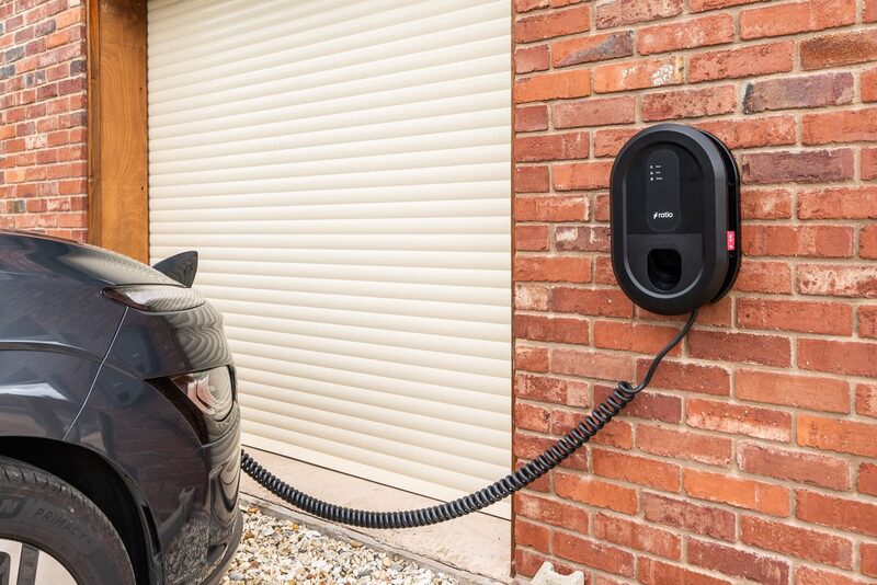 A black Ratio EV wall charger mounted on a red brick garage wall with a coiled cable plugged into a dark grey electric car