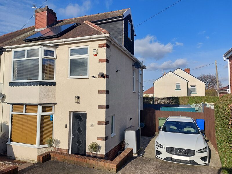 A UK semi-detached house with solar thermal panels on the roof and an air source heat pump unit beside the driveway, with a white Volvo parked outside
