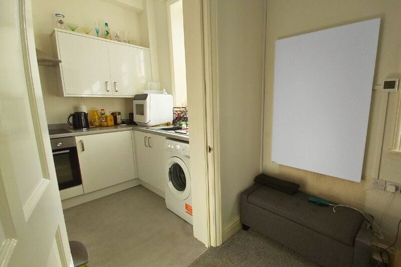 A white wall-mounted infrared heating panel installed in a small flat next to a kitchen with washing machine and oven