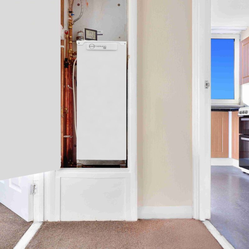 A Sunamp Thermin heat battery unit installed in an airing cupboard with copper pipework in a flat, with a kitchen visible through the doorway
