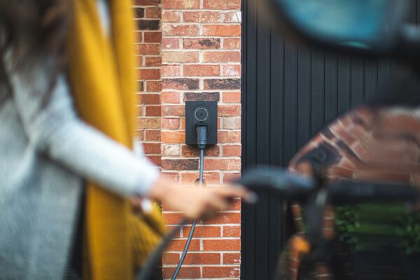 A wall-mounted EV charge point on a brick house wall with a person plugging the charging cable into a car