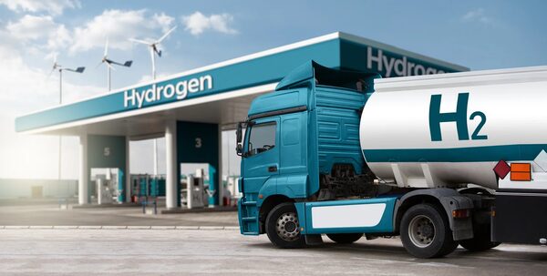A teal hydrogen tanker truck parked at a hydrogen refuelling station with wind turbines in the background