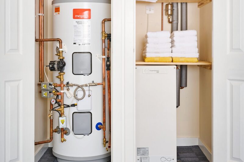 A Megaflo hot water cylinder with copper pipework in an airing cupboard next to a Sunamp heat battery unit and folded towels