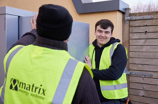 Two installers in hi-vis vests positioning an outdoor Viessmann Vitocal heat pump unit beside a house wall