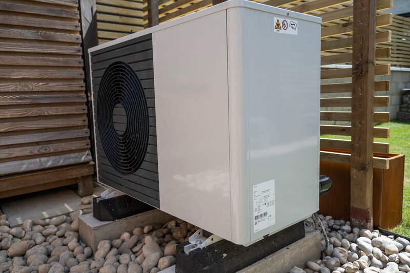 An air source heat pump unit installed outdoors on a concrete base among pebbles beside a wooden slatted fence