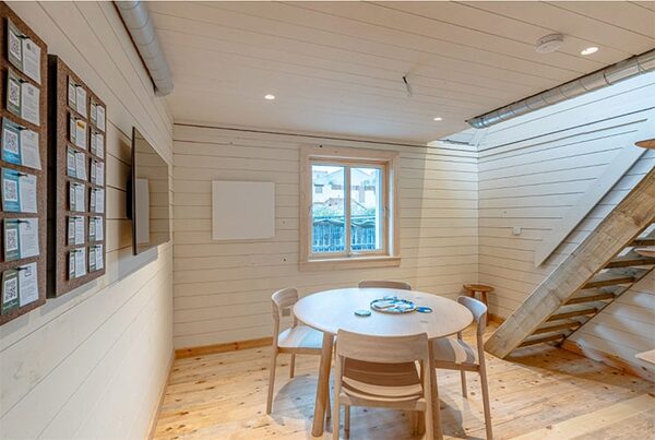 A white infrared heating panel mounted on the wall of a light timber-lined dining room with a round table, chairs and a wooden staircase
