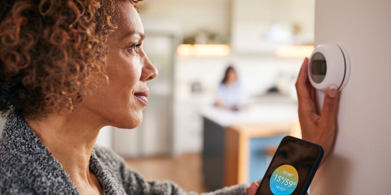 A woman adjusting a white smart thermostat on a wall while holding a smartphone showing a temperature app