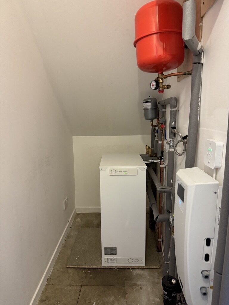 A Sunamp Thermino thermal battery hot water unit installed in a small airing cupboard with pipework, expansion vessel and red expansion tank