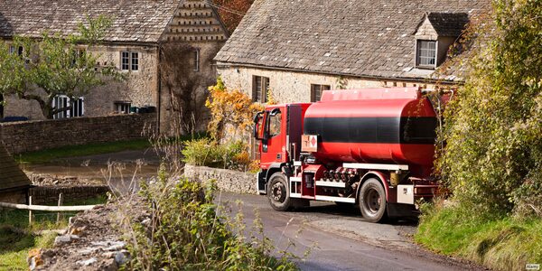 A red heating oil delivery tanker lorry driving along a lane past stone cottages in a village