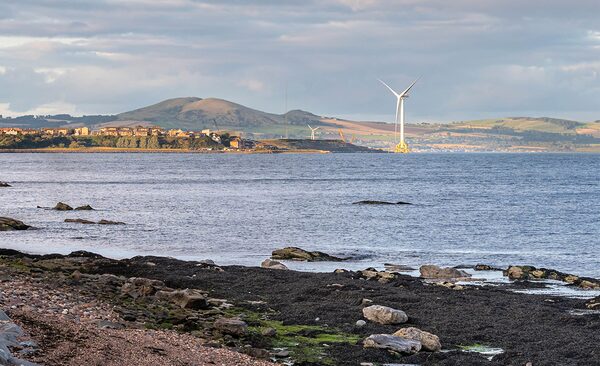 An offshore wind turbine in the sea near a Scottish coastline with hills and a rocky shore in the foreground