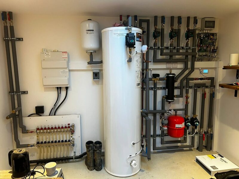 An installed heat pump system inside a home, with a large white hot water cylinder, expansion vessel, manifold, pipework, valves and controls on a white wall.