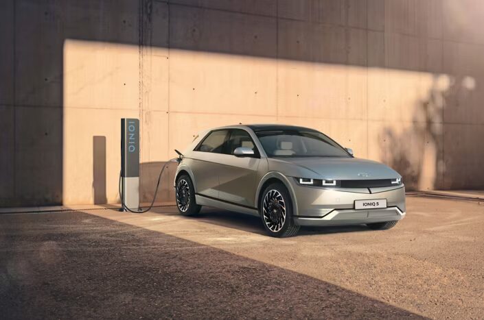 A silver Hyundai IONIQ 5 electric car plugged into an IONIQ-branded EV charger against a concrete wall