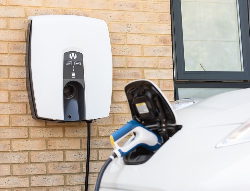 A white Indra wall-mounted EV charger on a brick house wall with a charging cable plugged into a white electric car